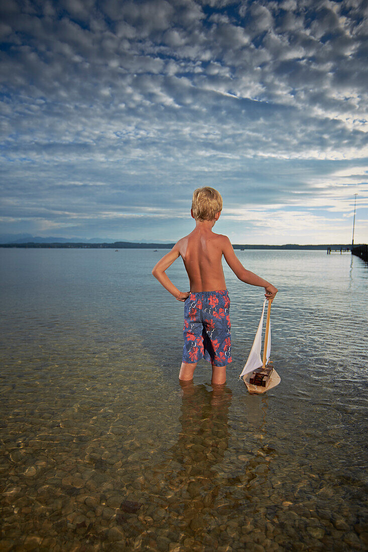 Boy with toy sail boat standing in lake Starnberg, Upper Bavaria, Bavaria, Germany