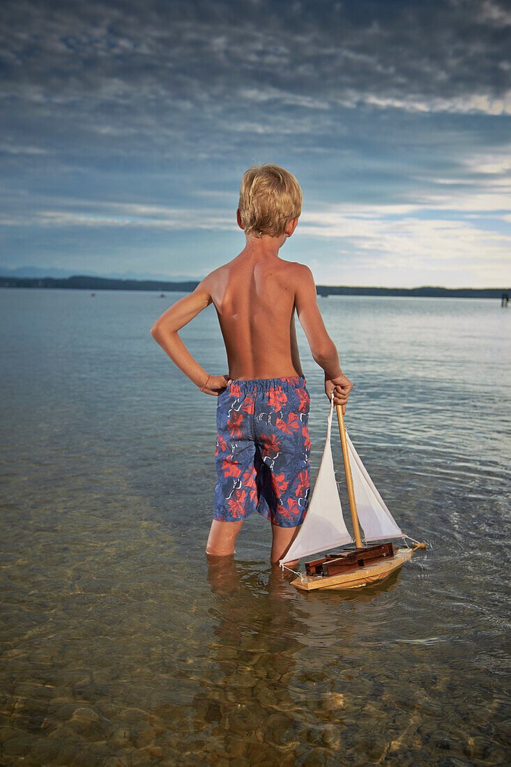 Boy with toy sail boat standing in lake Starnberg, Upper Bavaria, Bavaria, Germany