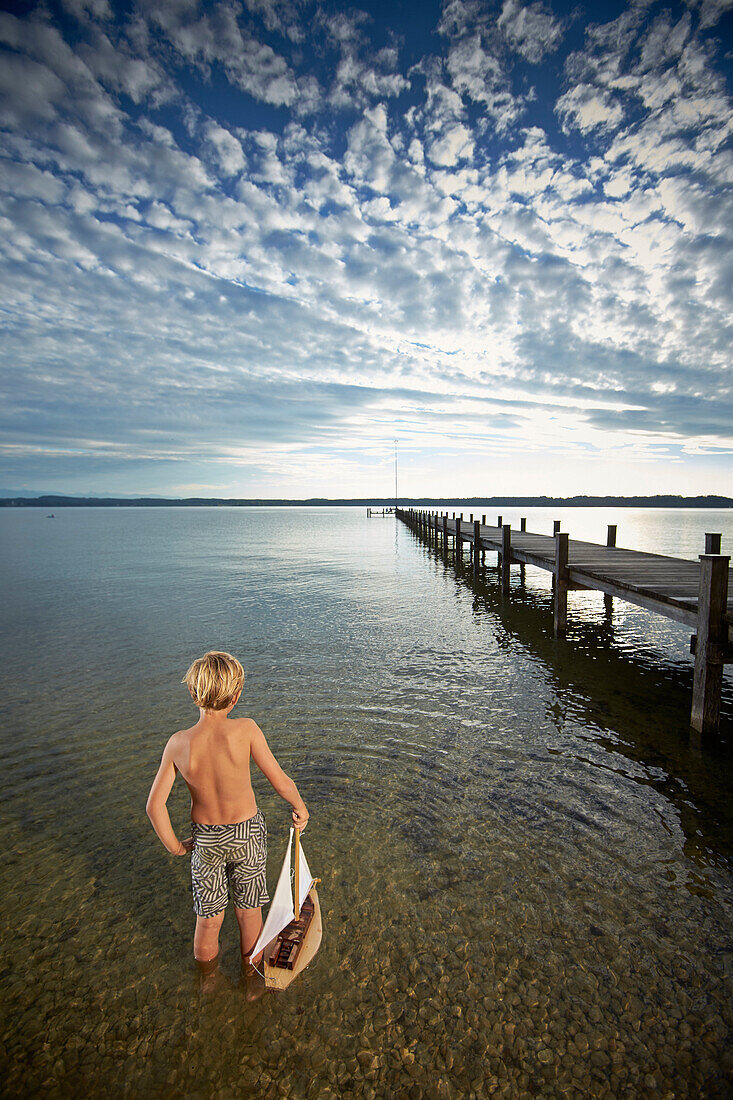 Boy with toy sail boat standing in lake Starnberg, Upper Bavaria, Bavaria, Germany