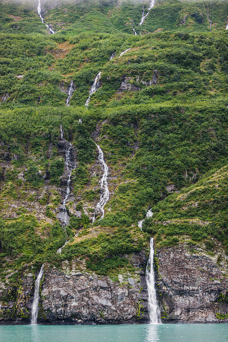 'Waterfalls spilling down a green forested mountain side over a cliff into Passage cannal, Prince William Sound; Whittier, Alaska, United States of America'