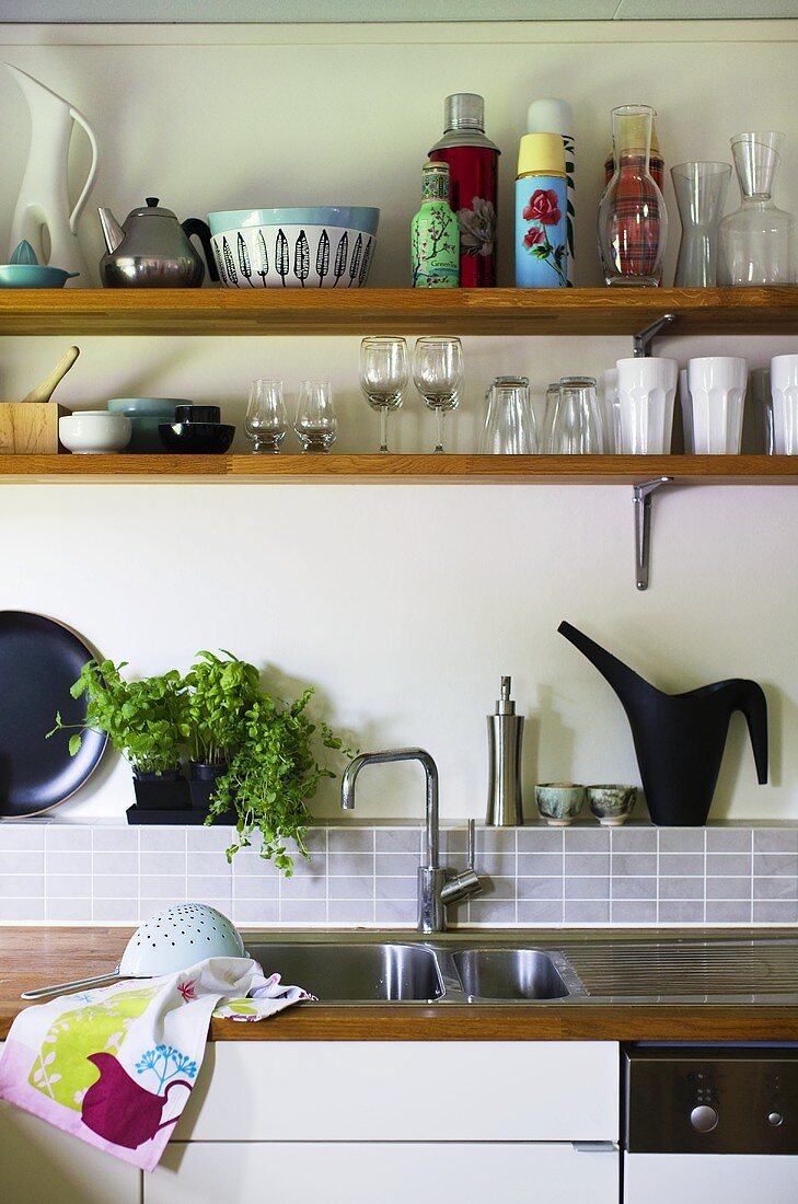 A kitchen sink with taps and a wooden shelf with glasses