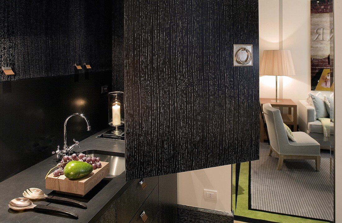 Open door of a black kitchen cabinet with a recessed sink in the counter and view of a living room