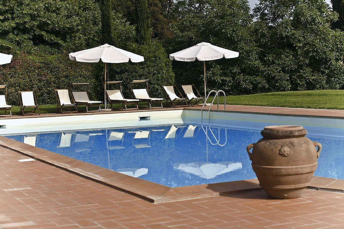 Clay planter beside a pool and lounge chairs with a sun umbrella reflected in the water