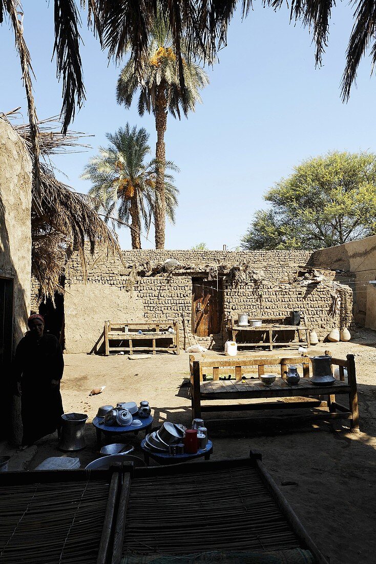 Bänke auf Sandboden im Innenhof mit Lehmmauern und hochgewachsenen Palmen, Ägypten
