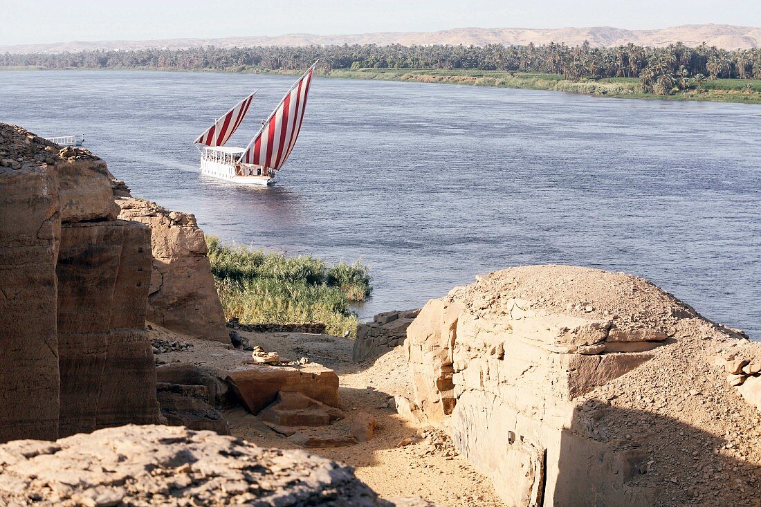Blick über Steinruinen auf Boot mit rotweissen Segeln auf dem Fluss, Nil, Ägypten