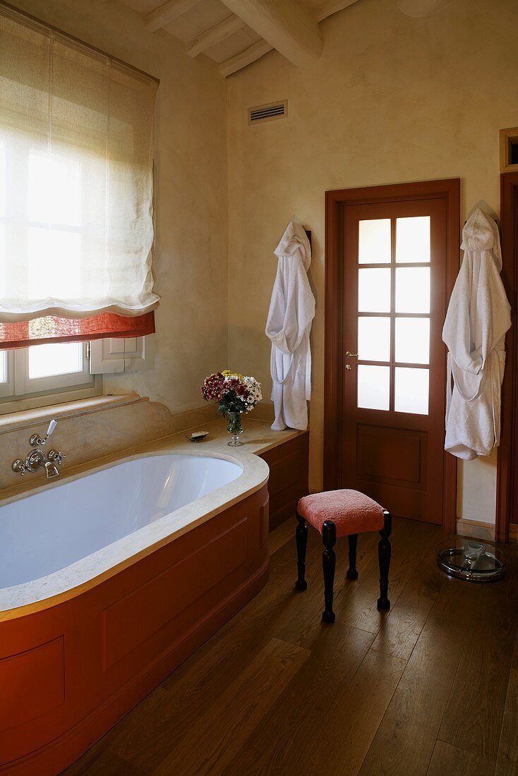 Ländliches Bad - Badewanne mit Holzverkleidung vor Fenster mit Rollo und gepolsterter Hocker auf Dielenboden