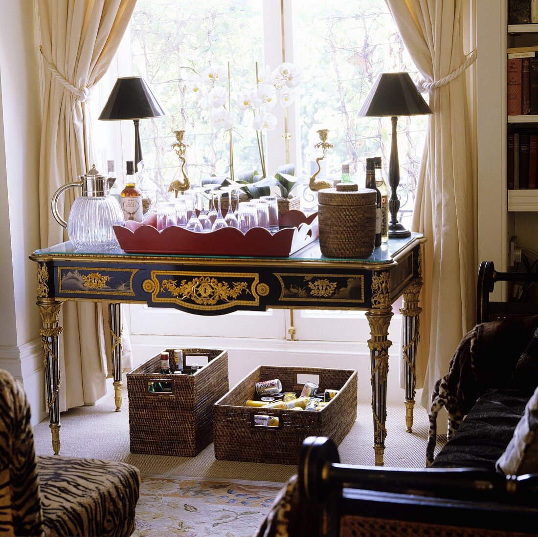 A tray of glasses on a Baroque table in front of floor-to-ceiling windows with bottles of alcohol underneath it in baskets