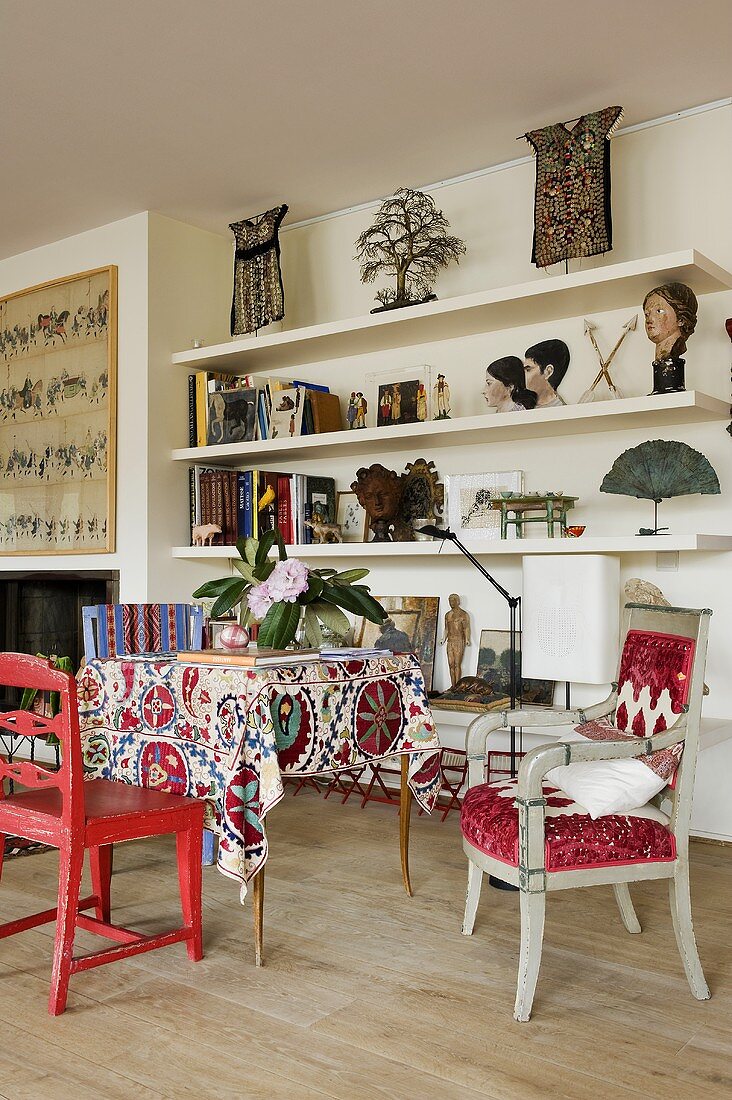 A living room with coloured chairs and a table with a colorful tablecloth in front of a white shelf wall