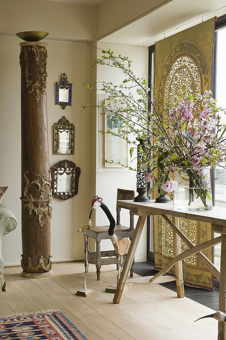 An ethnic style living room - antique wooden columns and a light wooden table in front of an ethnic cloth at a window