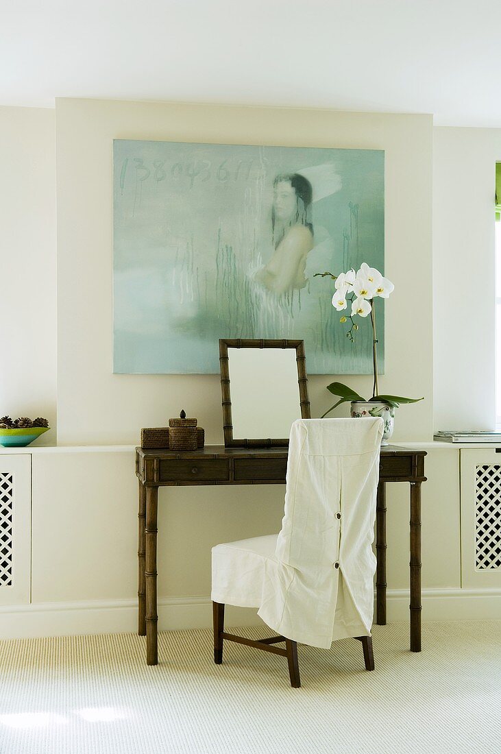 A bamboo table with a chair with a white cover against a wall hung with a picture