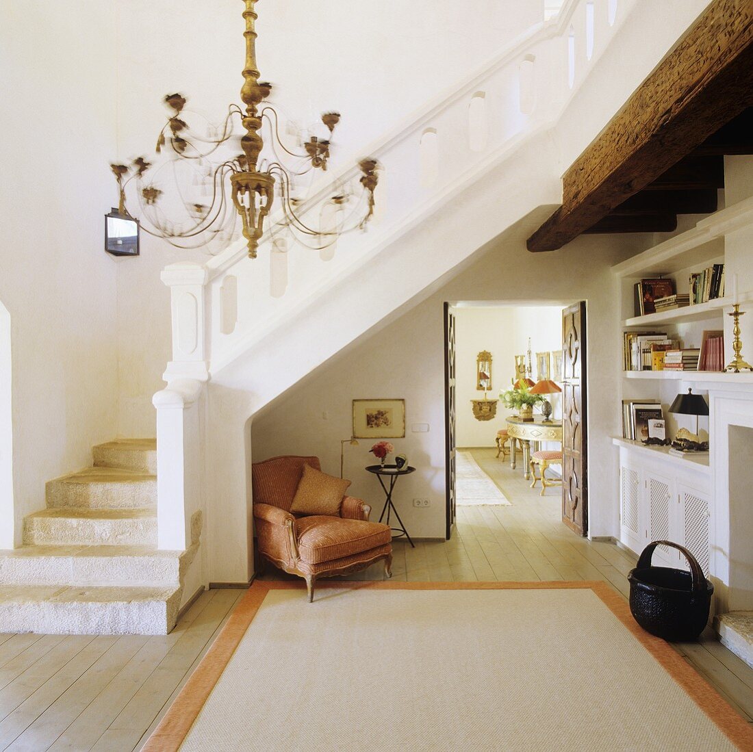 A spacious anteroom in a finca with a white-painted flight of stairs and a Baroque armchair under the stairs with a view through an open door