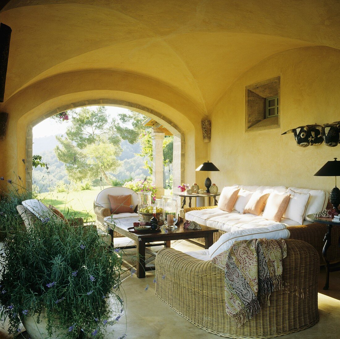 Gemütliche Korbmöbel mit weissen Polstern in gelbgetünchter Loggia mit Gartenblick