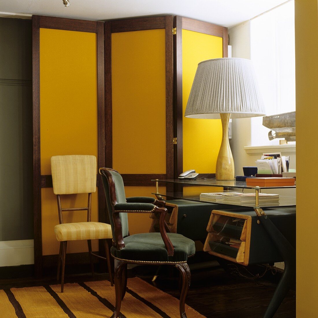 An antique desk chair in front of a glass table with a table lamp with a white pleated shade in front of a yellow paravent