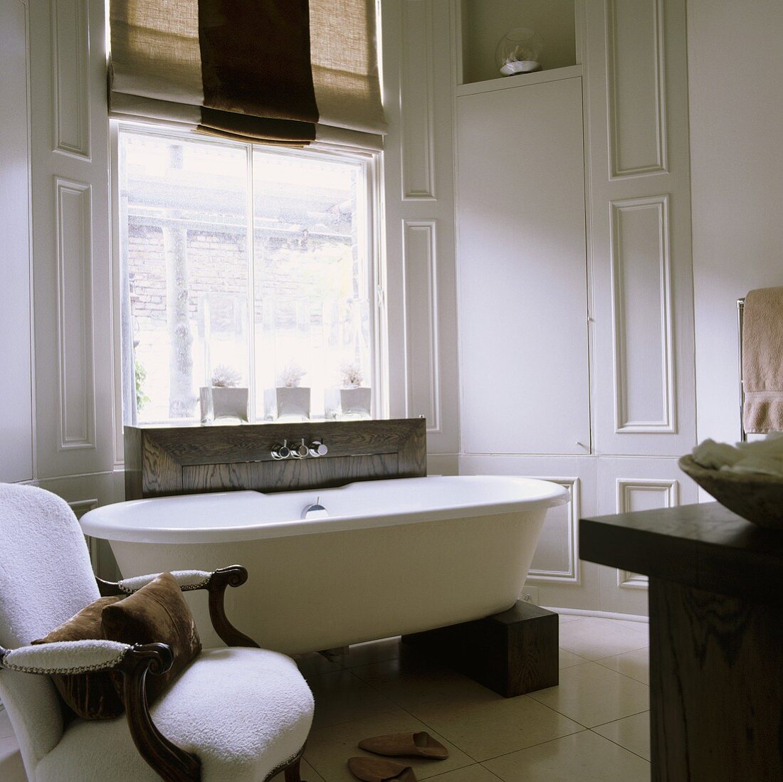 Freistehende Badewanne auf Holzklötzen und antiker weisser Sessel vor Fenster im hellgrau vertäfelten Bad