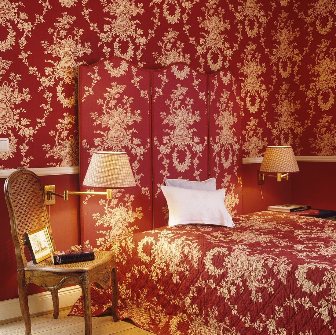 A bedroom with a red and white floral pattern on the wallpaper, the paravent and the bed cover