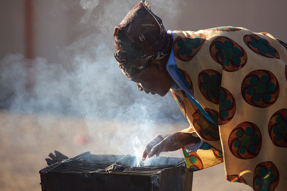 Herero Frau beugt sich über einen rauchenden Grill, Sesfontain, Namibia