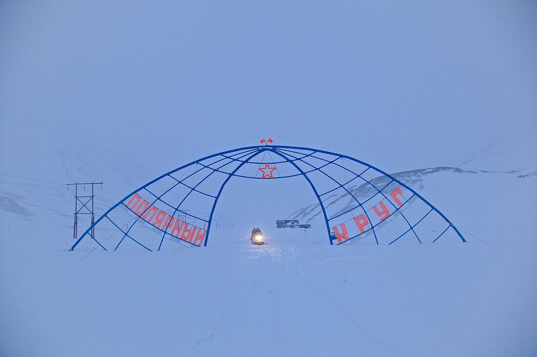 Snowmobil fährt im Schneetreiben durch Stahlbogendenkmal, das den Schnittpunkt des Nördlichen Polarkreises mit dem 180. Längengrad markiert, Tschukotka (Autonomer Kreis der Tschuktschen), Sibirien, Russland