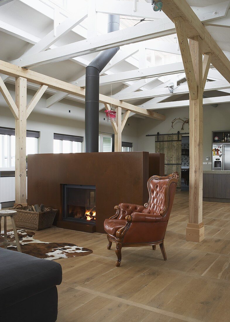 A rusty partition wall with a built-in fireplace and an antique leather armchair in front of wooden beams in an attic room