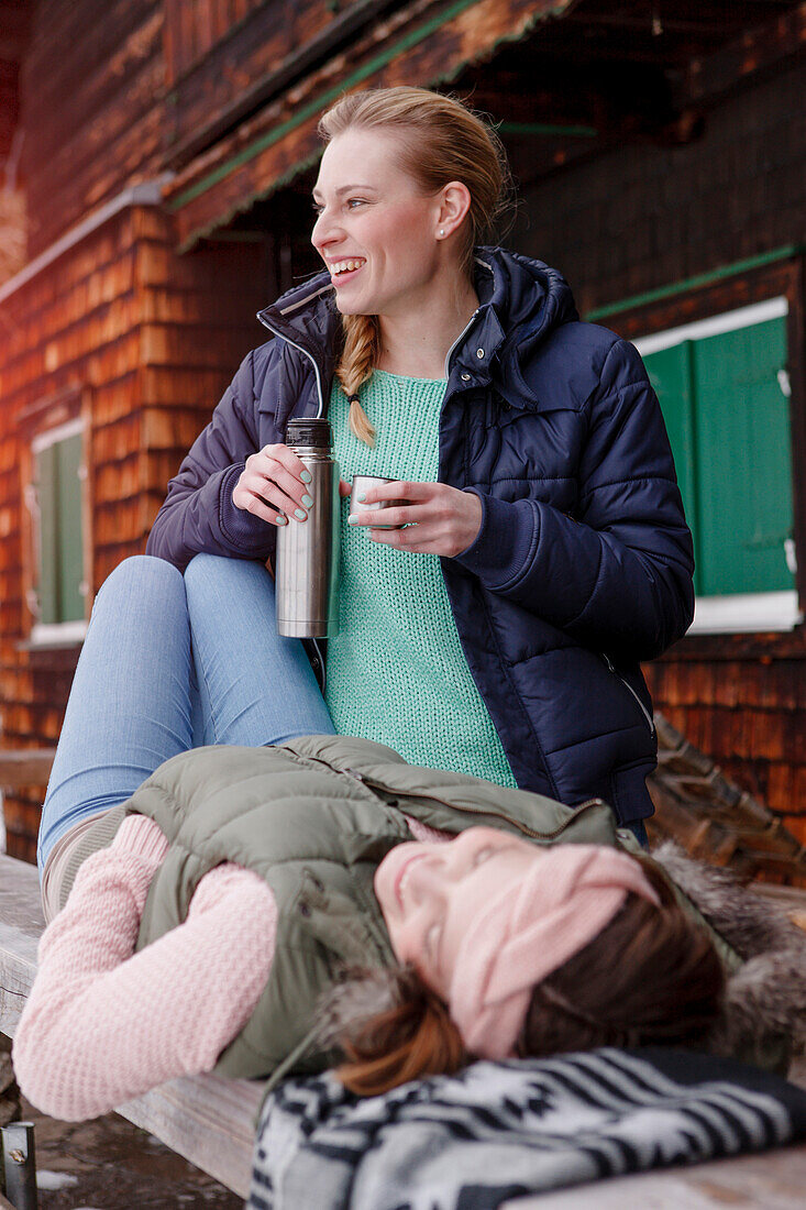 Two young women on a bench having a hot beverage, Spitzingsee, Upper Bavaria, Germany