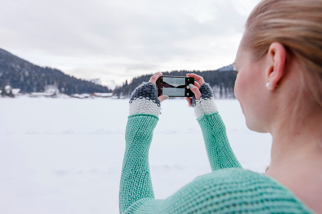 Junge Frau fotografiert mit einem Smartphone schneebedeckte Landschaft, Spitzingsee, Oberbayern, Bayern, Deutschland