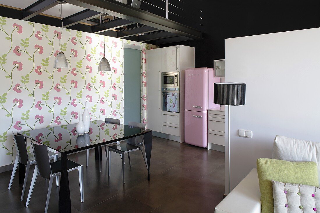 A living-room-cum-dining-room with a black shiny table against a wall hung with floral wall paper and a view into an open-plan kitchen