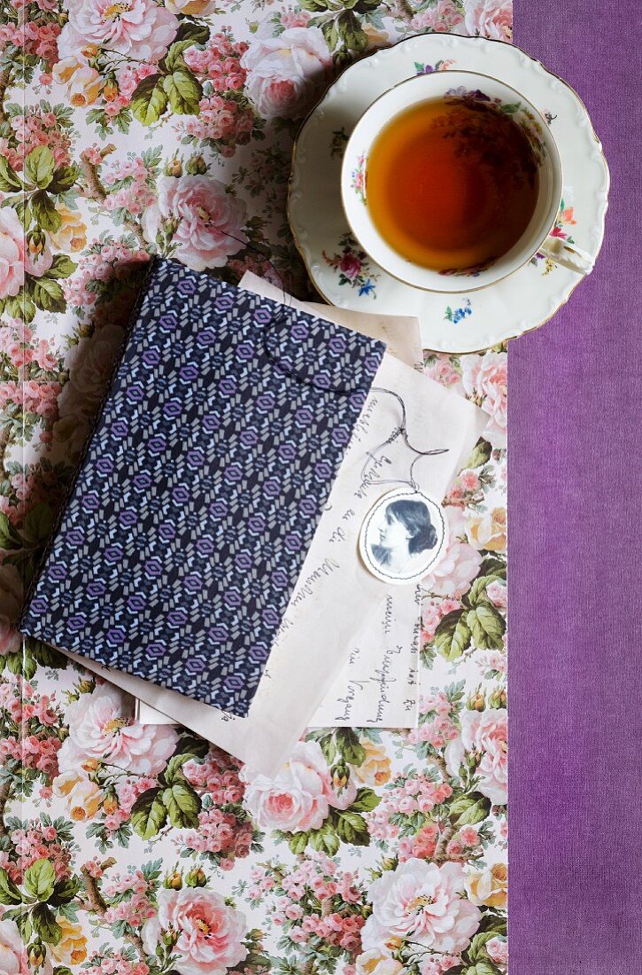 A cup of tea next to a book with a patterned cover on paper with a floral design