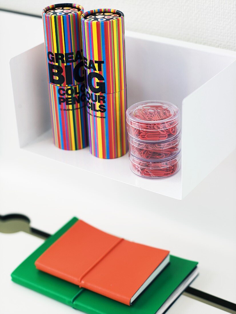 weiße Wandkonsole mit bunten Stiftboxen in Zylinderform und Hefte mit farbigem Einband
