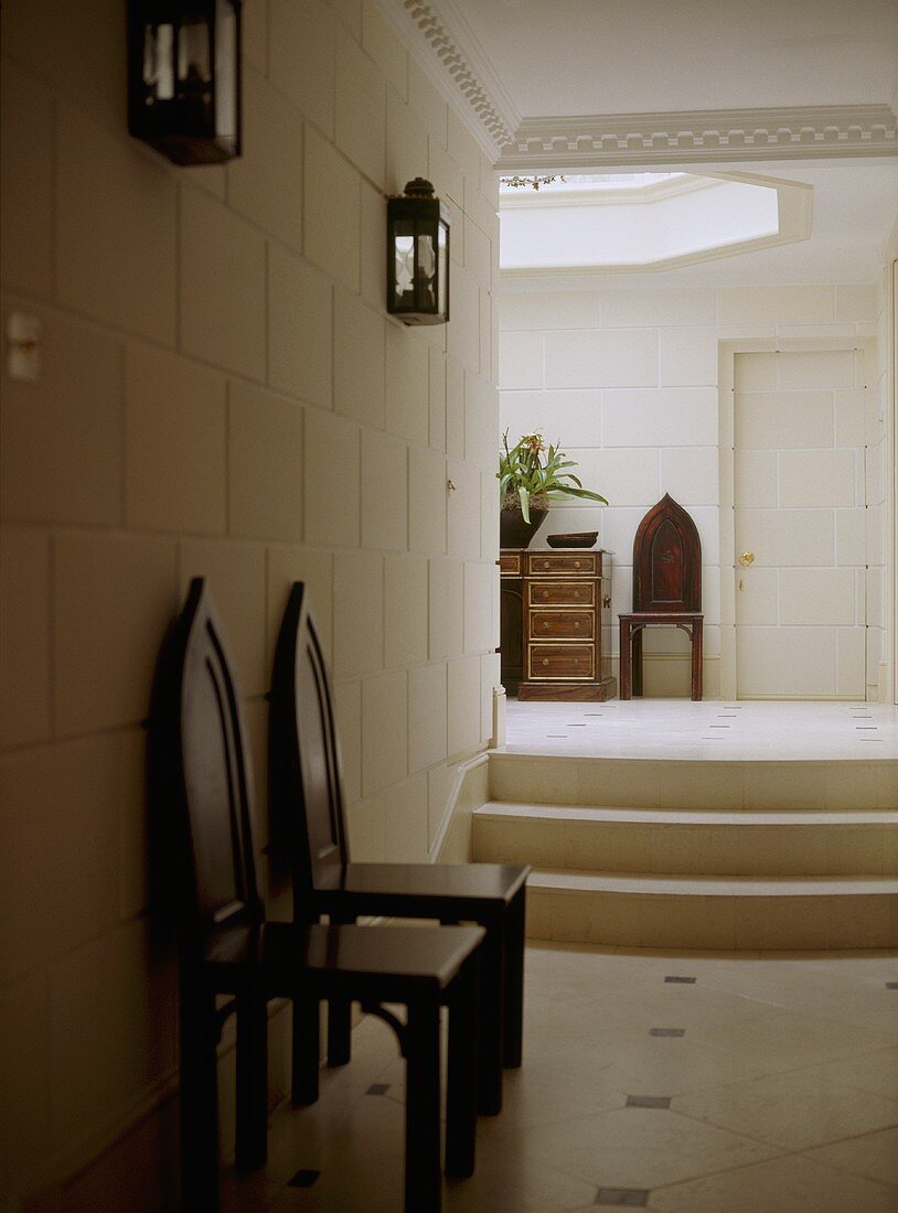 A modern hallway with steps leading up to higher level, a pair of wooden chairs with gothic style backs, tiled wall and floor