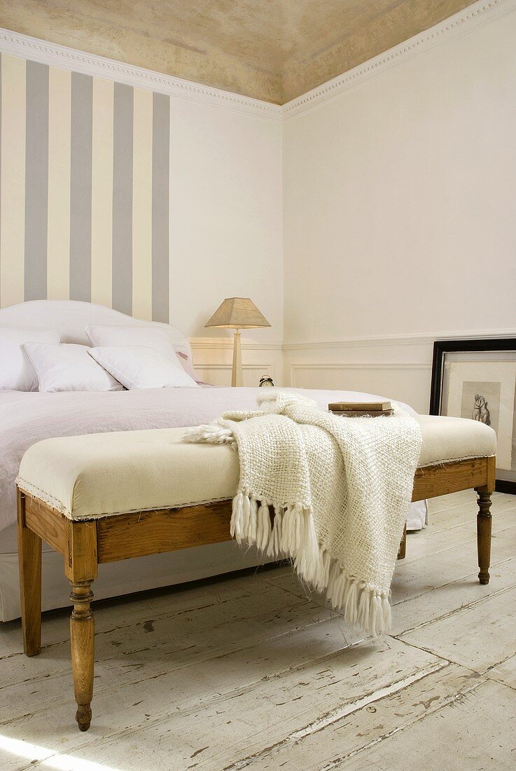 An antique bench with light-coloured upholstery and a knitted shawl at the end of a bed