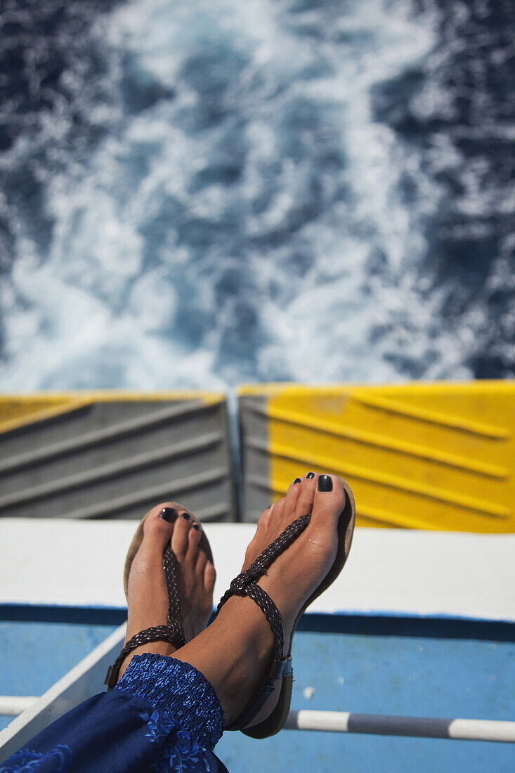 Füße einer Frau auf dem Sitz einer Deckfähre mit den Wellen im Hintergrund, Milos, Kykladen, Griechische Inseln, Griechenland, Europa.