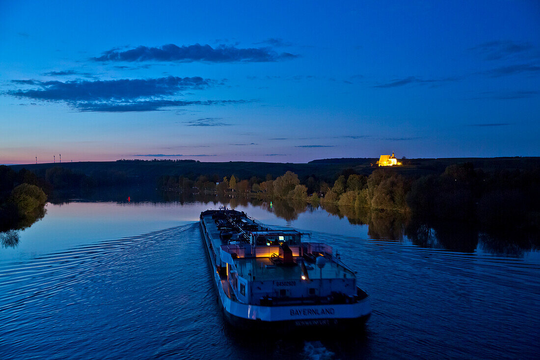 River freight ship Bayernland (Erik Walther GmbH & Co. KG) on the Main river with Maria im Weingarten pilgrimage church at dusk, Volkach, Franconia, Bavaria, Germany