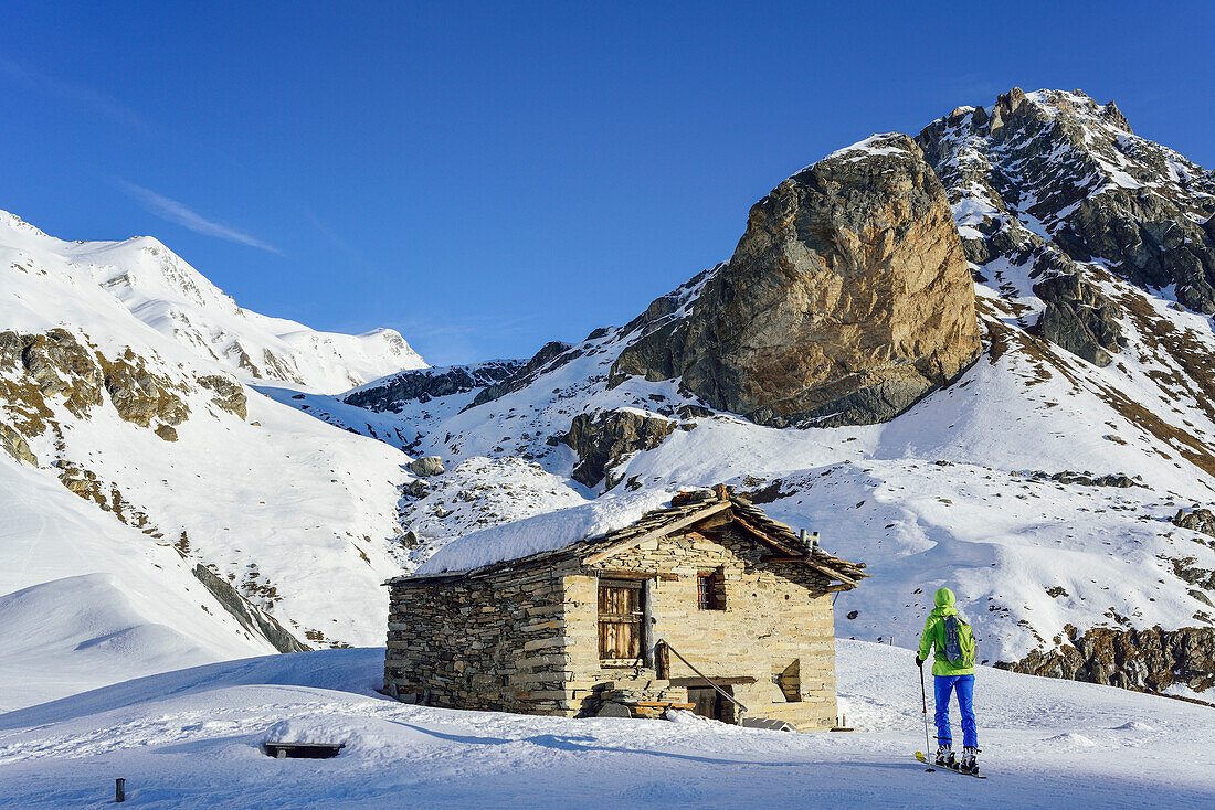 Frau auf Skitour steigt an Almhütte vorbei zum Monte Salza auf, Rocca Senghi im Hintergrund, Monte Salza, Valle Varaita, Cottische Alpen, Piemont, Italien