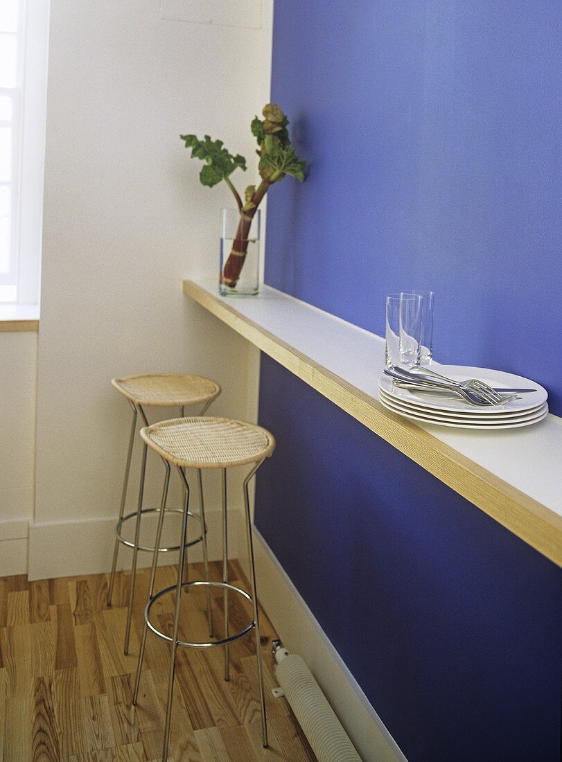 A detail of a modern blue kitchen, breakfast bar, stools, plates, cutlery, glasses, wooden flooring,