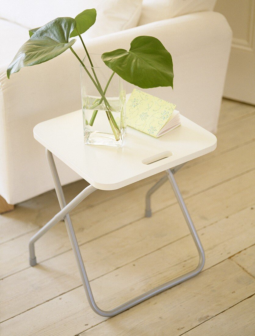 Glass vase of leaves on folding table next to white sofa.