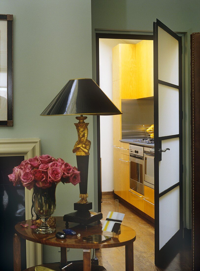 A detail of a traditional sitting room, door open with view into kitchen, side table, lamp, flower arrangement of pink roses,