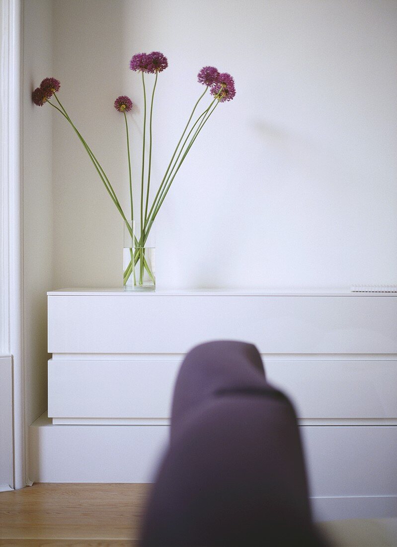 A detail of a modern, white bedroom, upholstered headboard in foreground, arrangement of Allium flowers in glass vase on unit,