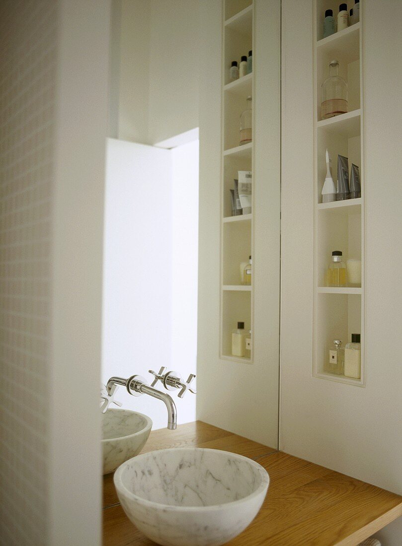 A detail of a modern bathroom in neutral colours, marble washbasin set on wooden shelf, built in shelving, taps, mirror, toiletries,