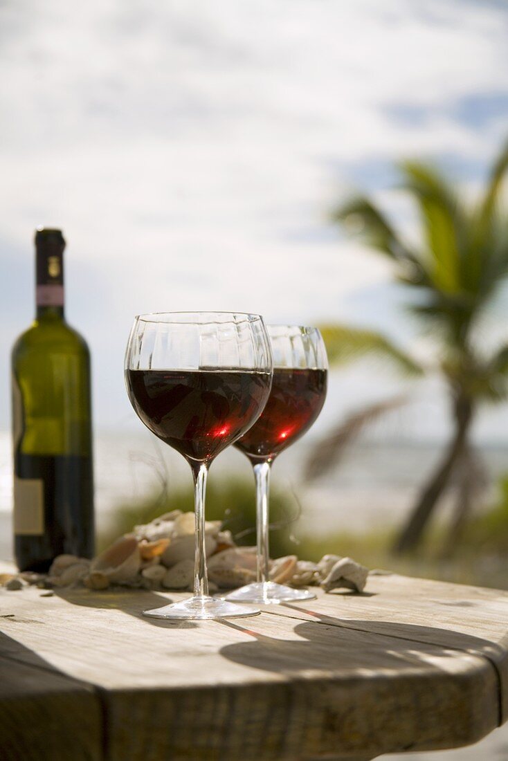 Zwei Rotweingläser mit Flasche auf Holztisch am Strand