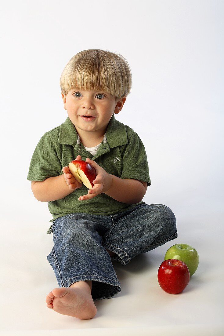 Ein sitzender kleiner Junge mit zwei ganzen und einem halben Apfel