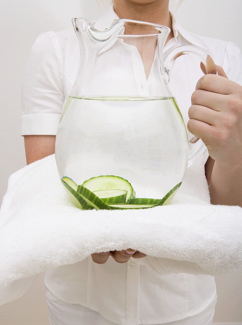 Frau hält Wasserkrug mit Gurkenscheiben auf Handtuch