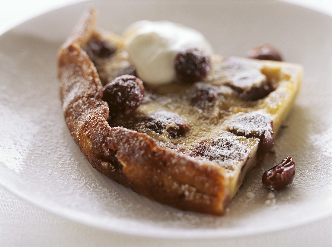 Ein Stück Clafouti mit Kirschen, Puderzucker und Sahne