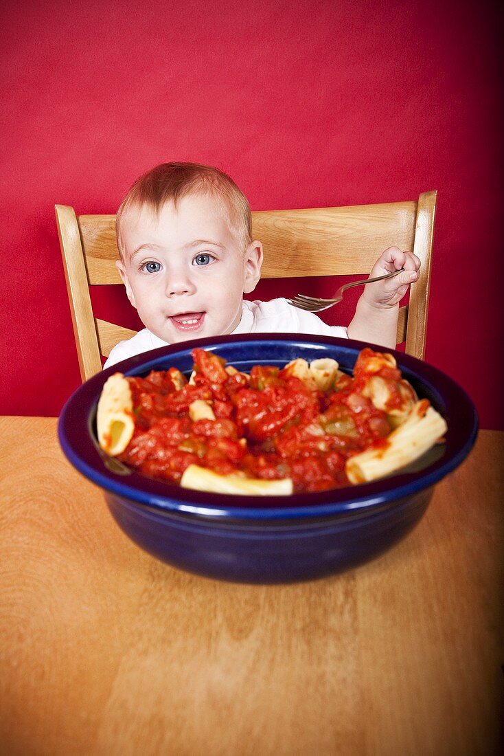 Baby at Table with Fork; Bowl of Pasta with Tomato Sauce on Table
