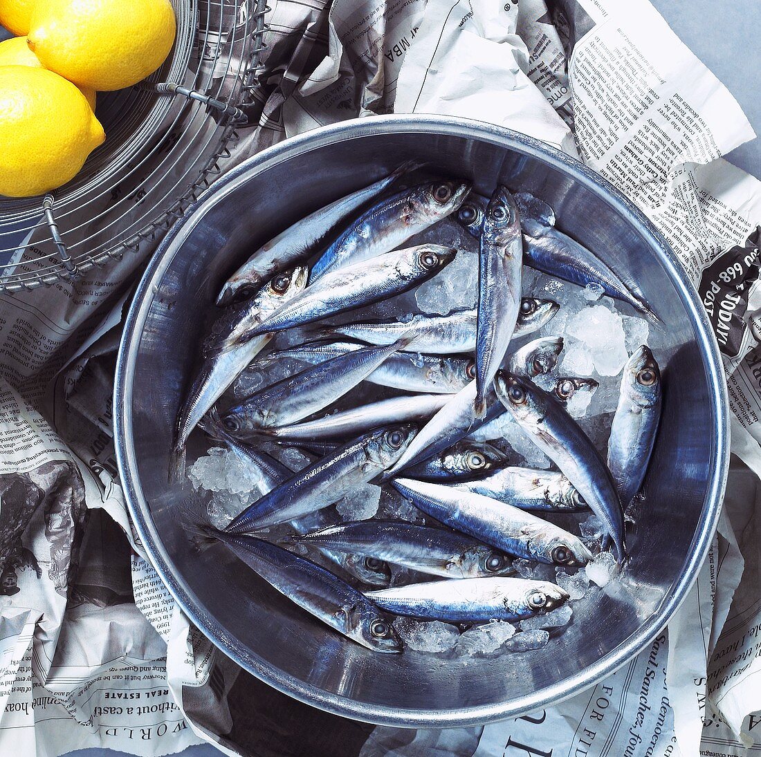 A bowl of fresh sardines on newspaper with lemons