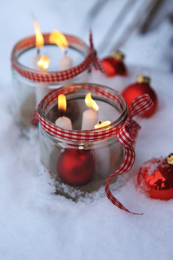 Einmachgläser mit brennenden Kerzen und Weihnachtskugeln im Schnee