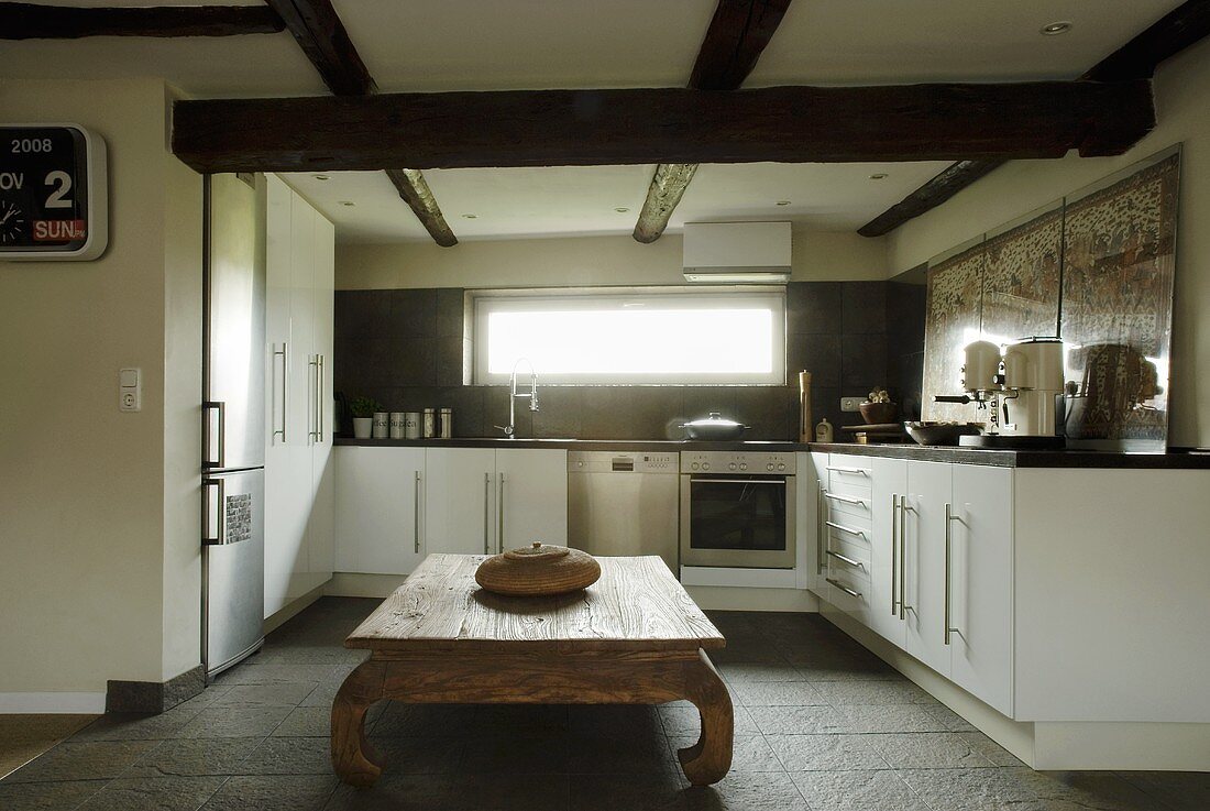 Offene Küche mit weissen Schrankfronten unter Holzbalkendecke und rustikalem Holztisch
