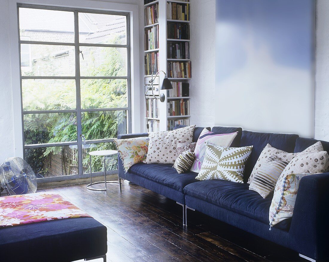 A purple sofa with cushions next to a bookshelf and a floor-to-ceiling window