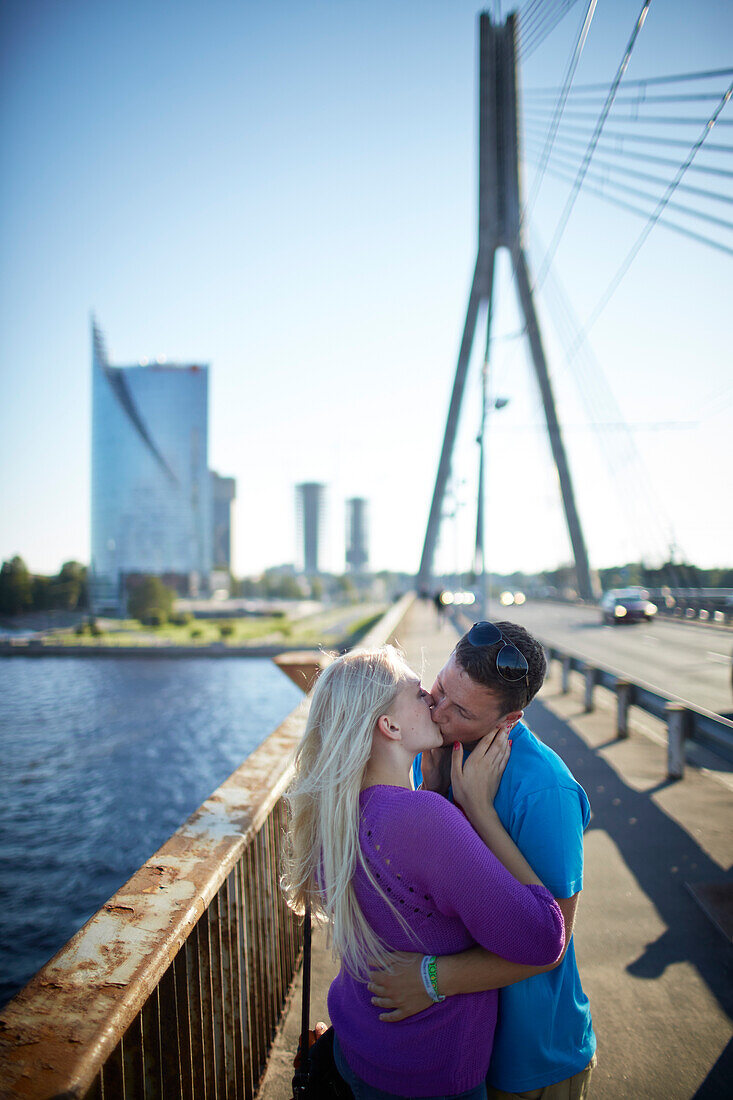 Couple on Vansu Tilts bridge, Svedbank office building The Sun Stone in the background, Daugava river, view towards Kipsala, Riga, Latvia