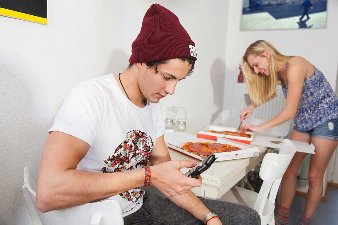 Young couple in the kitchen, playing with mobile and eating pizza