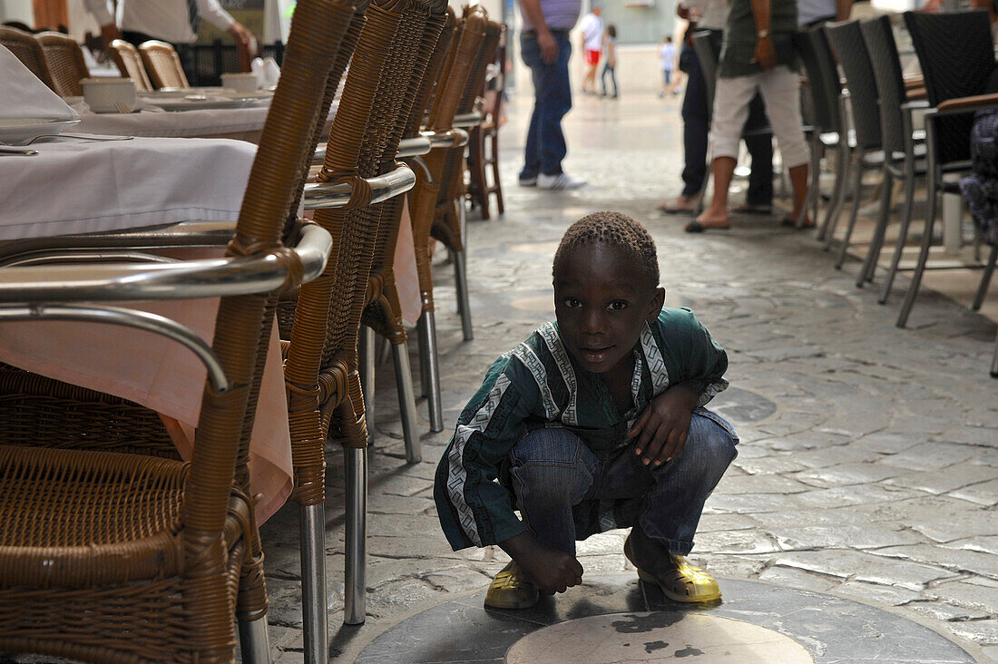 Black boy, child of a street vendor, between chairs of street bars at de Don Juan Diaz, near the Cathedral, Malaga, Andalusia, Spain, Europe