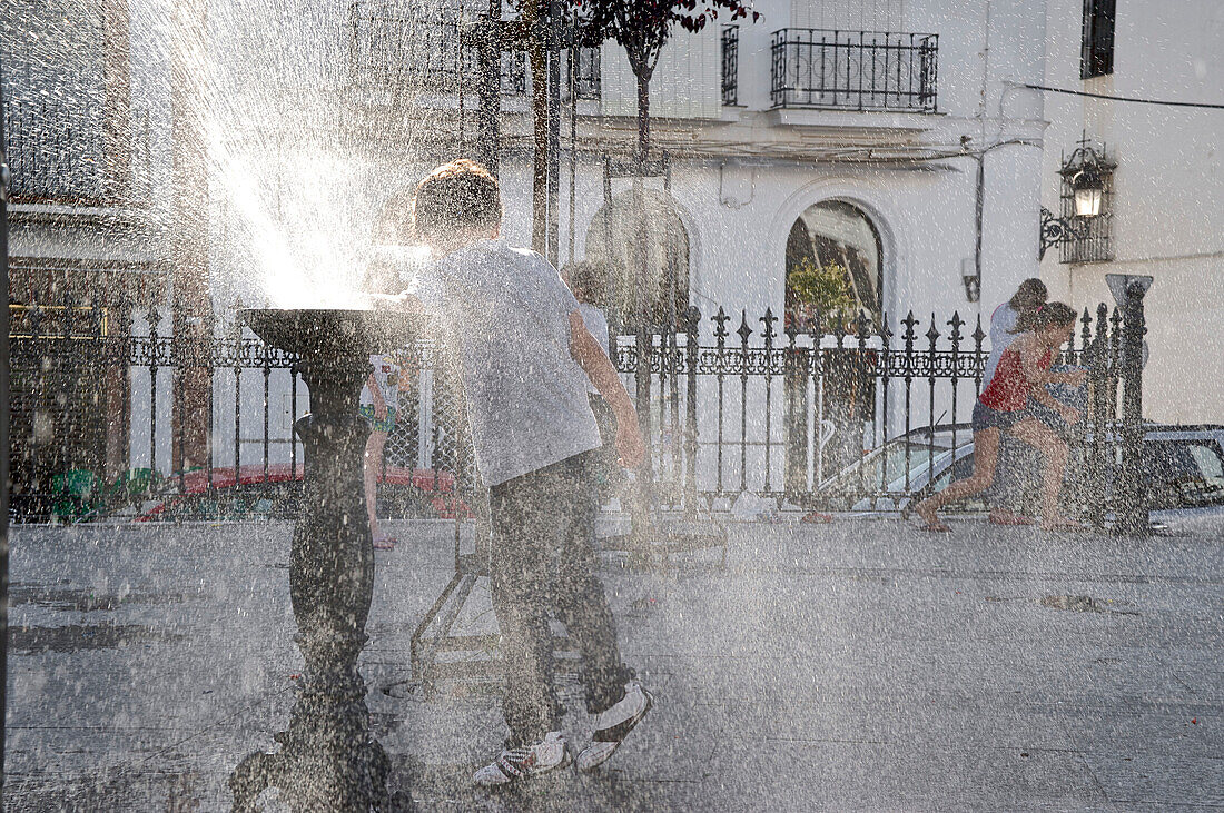 Children playing at a fountain and splashing with water, illuminated in the backlight, in the old town of Aracena, Huelva, Andalusia, Spain
